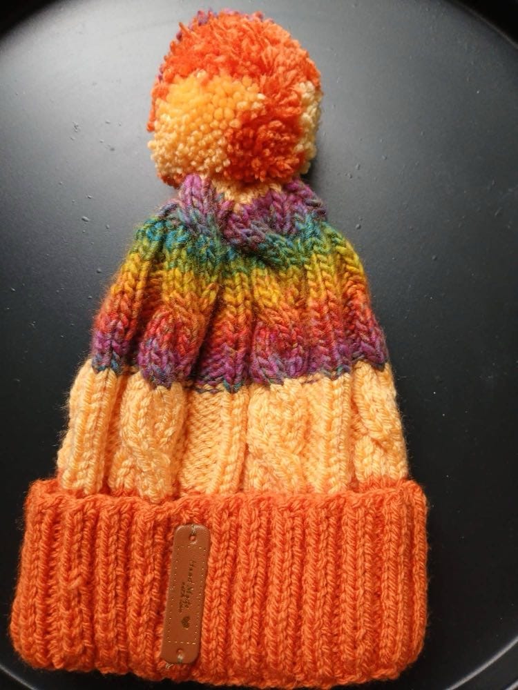 Cable-knit bobble hat with an orange cuff, peach body and multicoloured crown, and an orange and peach bobble